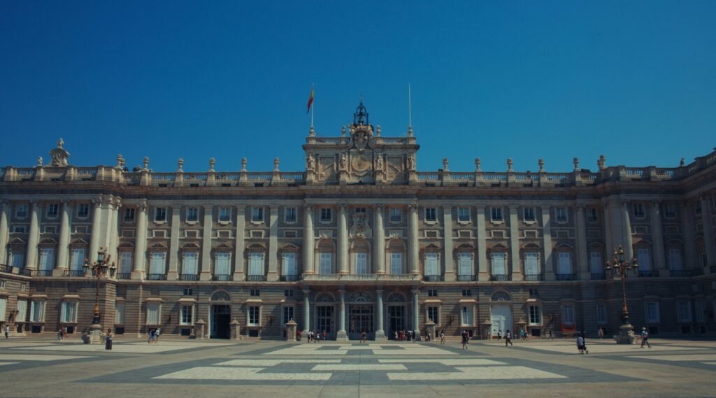 Royal Palace of Madrid, Spain, with its grand neoclassical facade and visitors walking in the main square under a clear blue sky. Madrid Winter Travel Guide 2025: Best Things to Do, See & Eat.